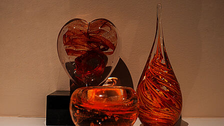 Drie glazen urnen van doorzichtig glas met oranje en rood, elk in een andere vorm. Een hart, een druppel en een bolvormige urn waar een waxinelichtje in kan.