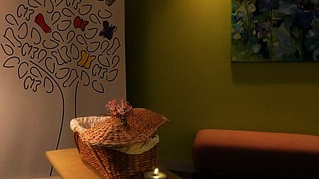 Een ruimte met een lichtgroene achterwand, een bankje en een lage tafel. Op een kunstwerk staat een boom afgebeeld in zwart op wit met enkele kleurrijke vlinders.