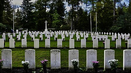 Nette rijen met grafstenen staan op een groen veld bij een monument in de vorm van een groot, wit kruis.