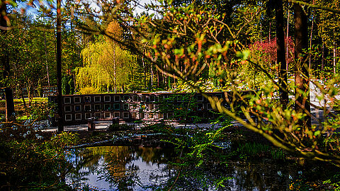 In een groene omgeving met bomen en water staat langs een pad een urnenmuur.