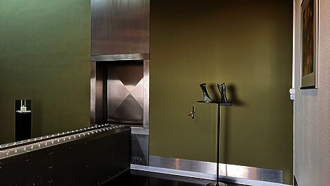 In de crematieruimte, met in het midden de invoerband voor de ovendeur met links daarvan een kaarsenstandaard en rechts van de oven een staand kunstwerk en een schilderij aan de muur.